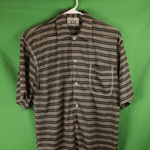 May include: A striped short-sleeved button-down shirt with a pocket on the left side. The shirt is brown and gray with a white star logo that says "All-Star" on the left side of the collar.