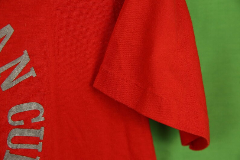 Op de afbeelding: Rood T-shirt met een zilveren afbeelding met de tekst "In Culture" aan de linkerkant.