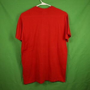 Op de afbeelding: Een effen rood T-shirt met korte mouwen. Het shirt hangt aan een witte hanger tegen een groene achtergrond.