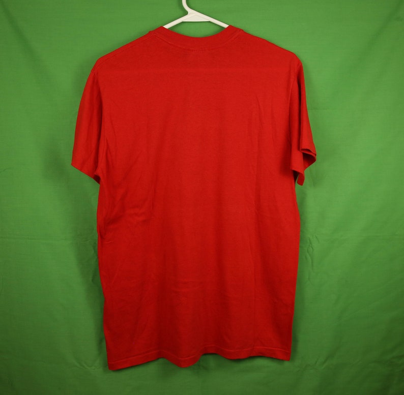 Op de afbeelding: Een effen rood T-shirt met korte mouwen. Het shirt hangt aan een witte hanger tegen een groene achtergrond.