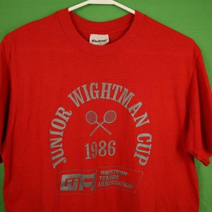 Op de afbeelding: Rood T-shirt met een zilveren print van het logo "Junior Wightman Cup 1986". Het logo toont twee gekruiste tennisrackets en de tekst "Western Tennis Association" in een kader.
