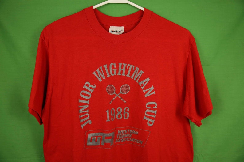 Op de afbeelding: Rood T-shirt met een zilveren print van het logo "Junior Wightman Cup 1986". Het logo toont twee gekruiste tennisrackets en de tekst "Western Tennis Association" in een kader.