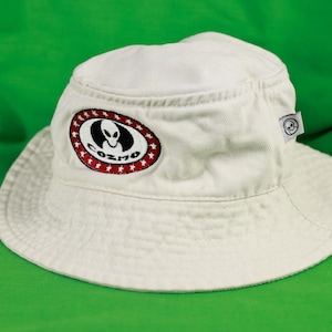 May include: White bucket hat with a wide brim. The hat features an embroidered patch with an alien design and the word "COZMO" in a red and white oval. A small tag with a skull logo is attached to the side.