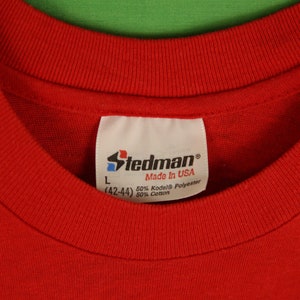 Op de afbeelding: Rood Stedman-merk t-shirt met een wit label met de tekst "Stedman Made in USA 50% Kodel&reg; Polyester (42-44) 50% Katoen"