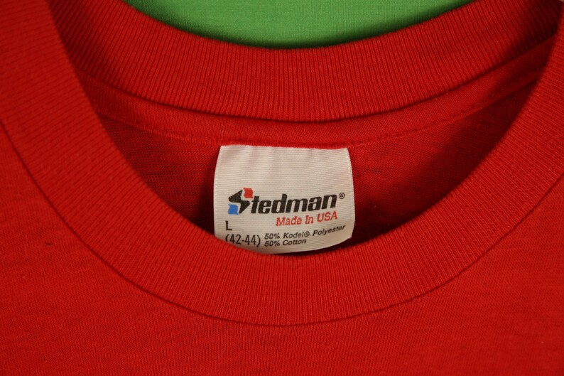 Op de afbeelding: Rood Stedman-merk t-shirt met een wit label met de tekst "Stedman Made in USA 50% Kodel&reg; Polyester (42-44) 50% Katoen"