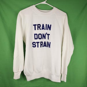 Peut inclure: Un sweat-shirt blanc avec le texte bleu marine "TRAIN DON'T STRAIN". Le haut à manches longues et col rond est suspendu à un cintre blanc sur un fond vert. Le sweat-shirt est en matière souple.