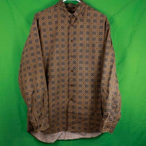 May include: A button-down shirt with a geometric pattern in shades of brown, tan, and gray. The long-sleeved shirt is hanging on a white hanger against a green background. The shirt has a collared neckline and buttoned cuffs.