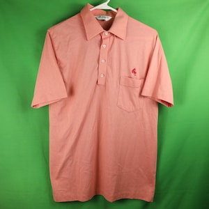 May include: A salmon pink short-sleeved polo shirt with a small embroidered logo on the left chest and a single pocket on the right side. The shirt has a button-down collar and a three-button placket.