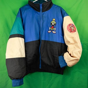 Puede incluir: Chaqueta con bloques de color azul, negro, turquesa y beige. La chaqueta tiene cremallera completa, cuello alto y un parche de personaje de dibujos animados en el pecho. La manga derecha tiene un parche circular con personajes de dibujos animados.