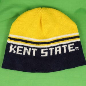 May include: A yellow and navy blue knitted beanie hat with the white text "KENT STATE". The hat features white and black stripes. The beanie is made of a soft, knitted material.