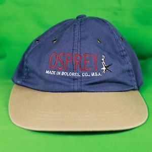 May include: A navy blue baseball cap with a beige peak. The word "OSPREY" is embroidered in red on the front, with a bird graphic. The text "MADE IN DOLORES, CO., U.S.A." is also embroidered on the cap.