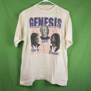 May include: Cream-colored t-shirt featuring the band name "GENESIS" in blue and purple lettering. The shirt displays three band member portraits and the text "We Can't Dance" in a stylized font. The design includes red and orange abstract shapes.