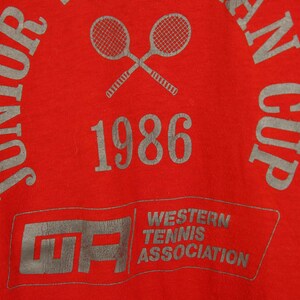 Op de afbeelding: Rood T-shirt met een grijze print van twee gekruiste tennisrackets, de tekst "Junior", "1986", "Western Tennis Association" en "An Cup".