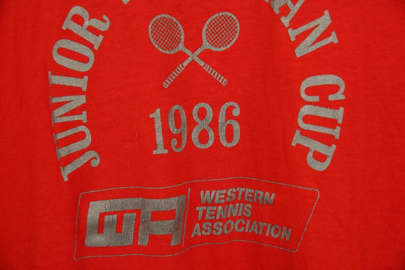 Op de afbeelding: Rood T-shirt met een grijze print van twee gekruiste tennisrackets, de tekst "Junior", "1986", "Western Tennis Association" en "An Cup".