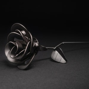 May include: A black metal rose with a single leaf, lying on a black surface.