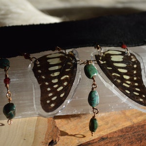 Könnte beinhalten: Eine Halskette mit zwei in klarem Harz gefassten Schmetterlingsflügeln, die an einem schwarzen Lederband hängen. Die Halskette hat türkisfarbene und rote Perlen an Kupferketten.