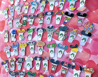 May include: A collection of 50 colorful enamel pins featuring various Disney characters and designs. Each pin depicts a stylized coffee cup with a different character or theme on top. The pins are arranged in rows on a pink and white background.