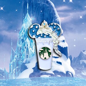 May include: A blue and white enamel pin featuring a stylized Mickey Mouse head with a snowflake design. The pin is topped with a crown and a figure of Elsa from the movie Frozen. The pin is in front of a blue and white background with a depiction of Elsa's ice castle.