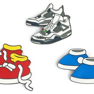 Puede incluir: Conjunto de cuatro pines de esmalte con diferentes diseños de zapatos. Los pines incluyen un par de zapatillas blancas y grises, un par de zapatillas rojas con detalles amarillos y un par de botines azules.