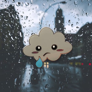 May include: A cartoon cloud with a sad face and a single teardrop. The cloud is a light beige color with a dark outline. The teardrop is a light blue color. The background is a blurry image of raindrops on a window.