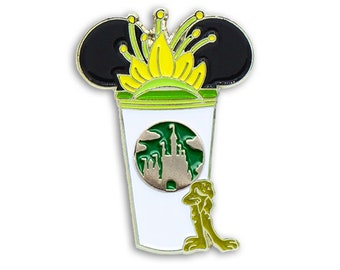 Può includere: Una tazza di caffè bianca con un coperchio verde e un cerchio verde con una silhouette di castello argentata. La tazza ha orecchie di Topolino nere con un fiore giallo in cima. Una rana verde è appoggiata alla tazza.
