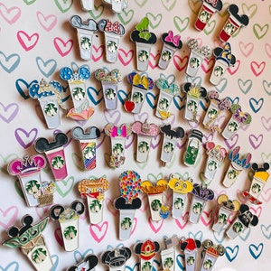 May include: A collection of 40 colourful enamel pins featuring various Disney characters and designs. Each pin depicts a Starbucks cup with a different character or theme, such as Mickey Mouse, Minnie Mouse, and Cinderella. The pins are arranged in rows on a pink and white background with hearts.