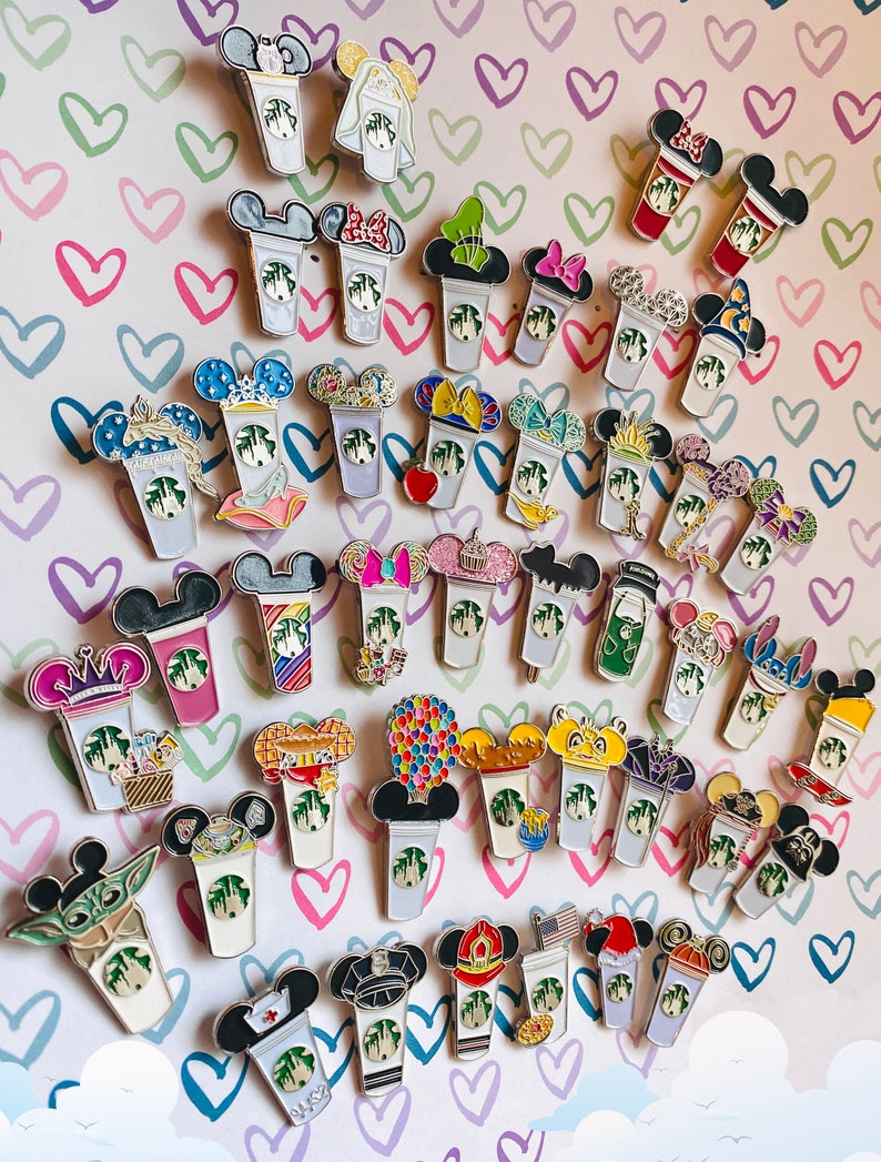 May include: A collection of 40 colourful enamel pins featuring various Disney characters and designs. Each pin depicts a Starbucks cup with a different character or theme, such as Mickey Mouse, Minnie Mouse, and Cinderella. The pins are arranged in rows on a pink and white background with hearts.