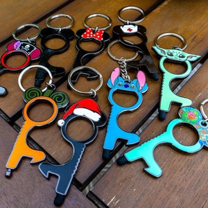 Könnte beinhalten: Eine Sammlung von zehn bunten Schlüsselanhängern mit verschiedenen Comicfiguren. Jeder Schlüsselanhänger hat ein kleines Metallwerkzeug, das zum Öffnen von Türen und Drücken von Knöpfen verwendet werden kann, ohne Oberflächen zu berühren.
