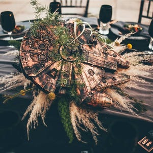 May include: A copper-colored model of the Millennium Falcon spaceship from Star Wars is surrounded by dried grasses and greenery, creating a unique centerpiece for a wedding or event.