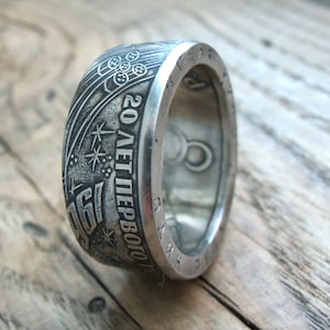 Puede incluir: Un anillo de plata hecho de una moneda con la inscripción "20 лет первого полета" (20 años del primer vuelo) y un diseño de estrellas y un cohete.