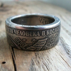 Op de afbeelding: Een zilveren ring met de inscriptie "50 лет человека в космосе" (50 jaar mens in de ruimte) en een afbeelding van een raket en sterren.