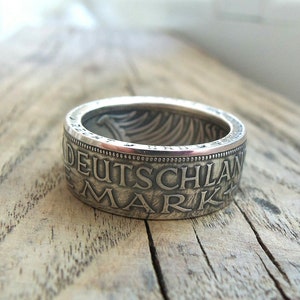 Può includere: Un anello d'argento realizzato con una moneta tedesca Mark. L'anello presenta l'iscrizione "DEUTSCHLAND MARK" attorno alla fascia.