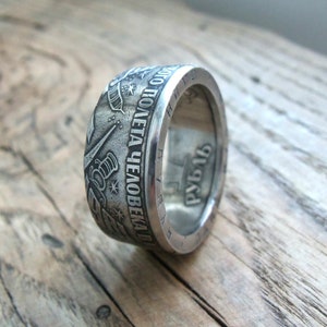 Op de afbeelding: Een zilveren ring gemaakt van een munt met de inscriptie "ВСЕГДА ПОЛЕТА ЧЕЛОВЕКА В КОСМОС" en het jaar "1961" aan de zijkant. De ring ligt op een houten oppervlak.
