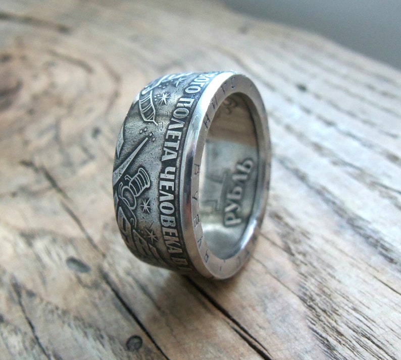 Op de afbeelding: Een zilveren ring gemaakt van een munt met de inscriptie "ВСЕГДА ПОЛЕТА ЧЕЛОВЕКА В КОСМОС" en het jaar "1961" aan de zijkant. De ring ligt op een houten oppervlak.