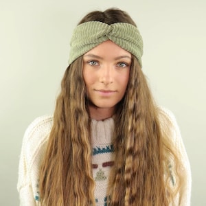 以下が含まれることがあります： 長い波状の茶色の髪をした女性が、緑色のコーデュロイのヘッドバンドをしています。ヘッドバンドはねじれていて、上に結び目があります。彼女はカラフルな模様の白いセーターを着ています。