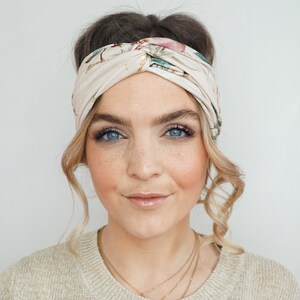 K&ouml;nnte beinhalten: Ein cremefarbenes Stirnband mit floralem Muster. Das Stirnband ist oben geknotet und wird um den Kopf getragen. Das florale Muster umfasst rosa, gr&uuml;ne und blaue Akzente. Die Person auf dem Bild tr&auml;gt einen beigen Pullover.