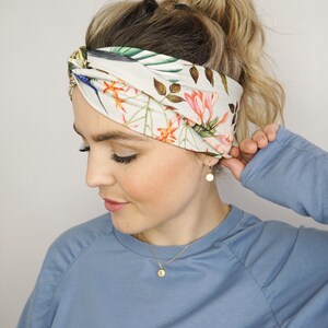 Puede incluir: Una diadema con estampado floral con fondo crema, hojas verdes, flores naranjas y rosas y detalles de p&aacute;jaros azules. La diadema retorcida se lleva alrededor de la cabeza. Tambi&eacute;n se ven pendientes y un collar dorados.