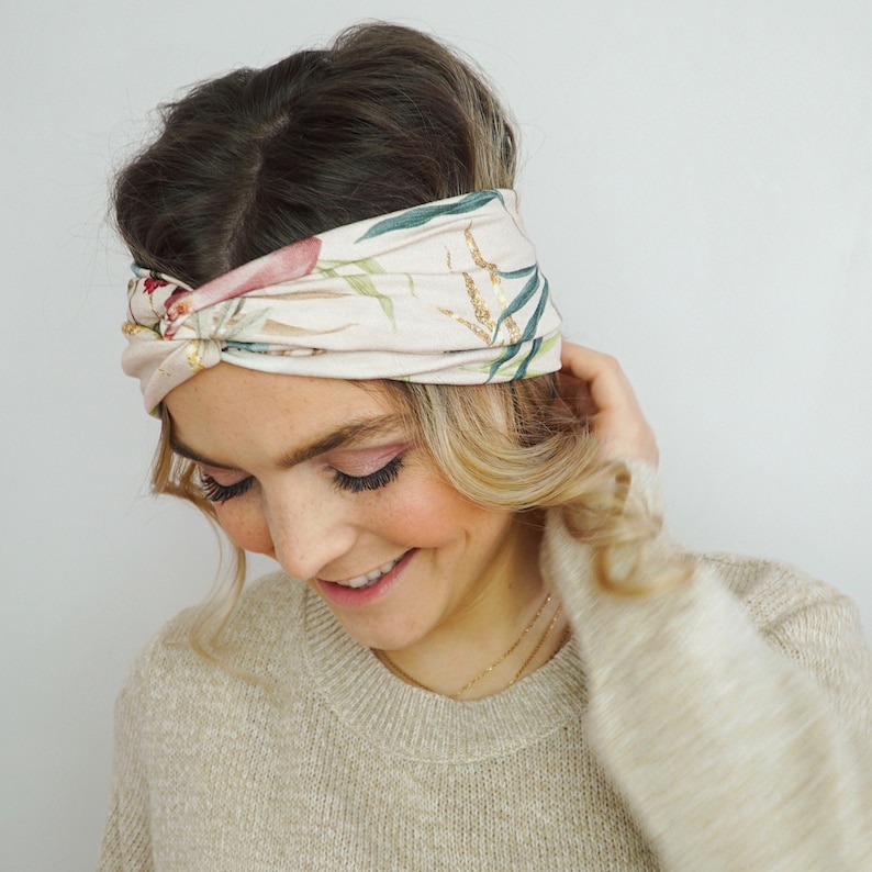 K&ouml;nnte beinhalten: Ein beiges Stoff-Haarband mit einem floralen Muster in Rosa, Gr&uuml;n und Gold. Das Haarband ist oben geknotet und wird um den Kopf getragen.