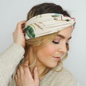 K&ouml;nnte beinhalten: Ein beiges Stirnband mit tropischem Blumenmuster mit gr&uuml;nen Bl&auml;ttern und roten und blauen Bl&uuml;ten. Das Stirnband ist um den Kopf gewickelt. Die Person tr&auml;gt einen beigen Pullover.