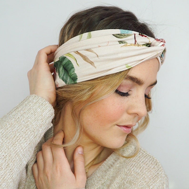 K&ouml;nnte beinhalten: Ein beiges Stirnband mit tropischem Blumenmuster mit gr&uuml;nen Bl&auml;ttern und roten und blauen Bl&uuml;ten. Das Stirnband ist um den Kopf gewickelt. Die Person tr&auml;gt einen beigen Pullover.