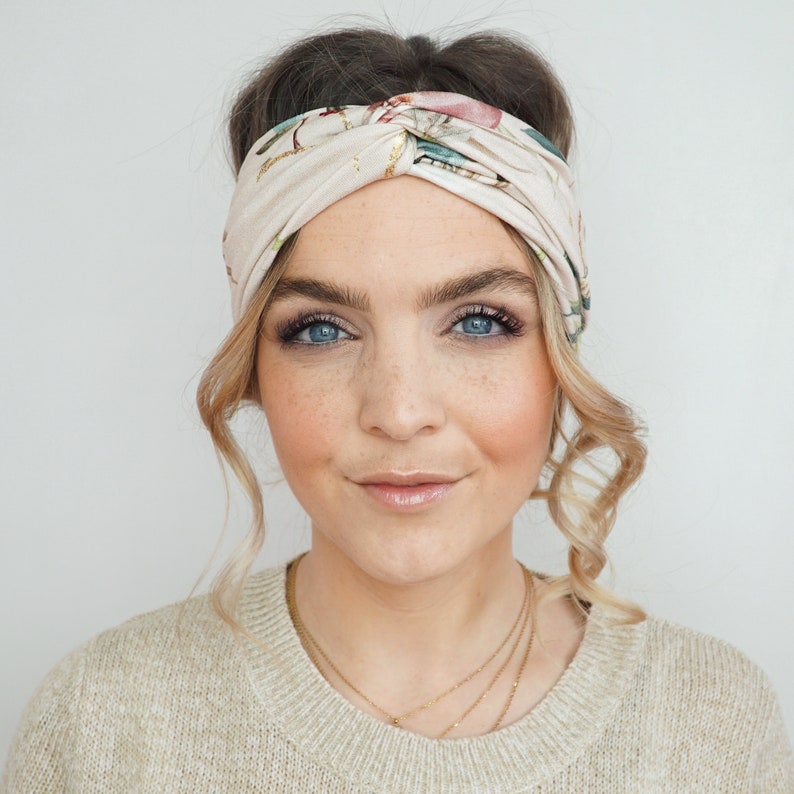 K&ouml;nnte beinhalten: Ein cremefarbenes Stirnband mit floralem Muster. Das Stirnband ist oben geknotet und wird um den Kopf getragen. Das florale Muster umfasst rosa, gr&uuml;ne und blaue Akzente. Die Person auf dem Bild tr&auml;gt einen beigen Pullover.