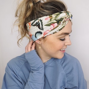 Puede incluir: Una diadema de color crema con estampado de mariposas y flores. La diadema presenta un patr&oacute;n repetido de mariposas en tonos negros, azules y naranjas, junto con elementos florales rosas y verdes. La tela est&aacute; envuelta y anudada en la parte superior.