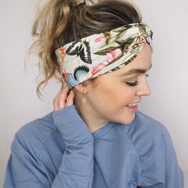 Puede incluir: Una diadema de color crema con estampado de mariposas y flores. La diadema presenta un patr&oacute;n repetido de mariposas en tonos negros, azules y naranjas, junto con elementos florales rosas y verdes. La tela est&aacute; envuelta y anudada en la parte superior.