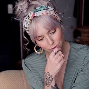 Puede incluir: Una diadema con estampado floral con detalles en verde, rosa y blanco. La persona lleva pendientes de aro dorados y varios anillos dorados. Un tatuaje de mandala es visible en la mu&ntilde;eca. La diadema est&aacute; envuelta alrededor de la cabeza.