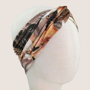 Op de afbeelding: Een hoofdband met bloemenprint in bruin-, beige- en oranjetinten wordt getoond op een mannequinhoofd. De stof is aan de bovenkant gedraaid, waardoor een stijlvol accessoire ontstaat. De hoofdband is ontworpen om om het hoofd te worden gedragen.