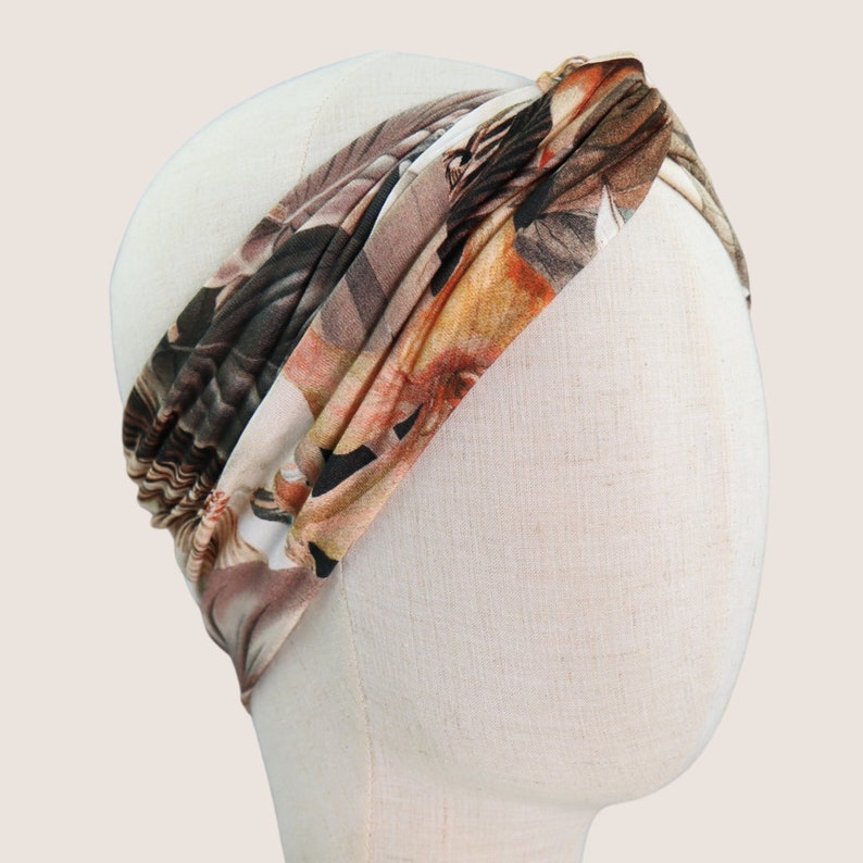 Op de afbeelding: Een hoofdband met bloemenprint in bruin-, beige- en oranjetinten wordt getoond op een mannequinhoofd. De stof is aan de bovenkant gedraaid, waardoor een stijlvol accessoire ontstaat. De hoofdband is ontworpen om om het hoofd te worden gedragen.