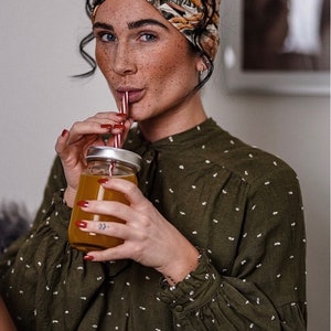 Op de afbeelding: Een vrouw met een patroon hoofdband en een groene blouse met witte stippen drinkt uit een glazen pot met een rietje. De pot bevat een oranje vloeistof. De vrouw heeft rode nagellak.