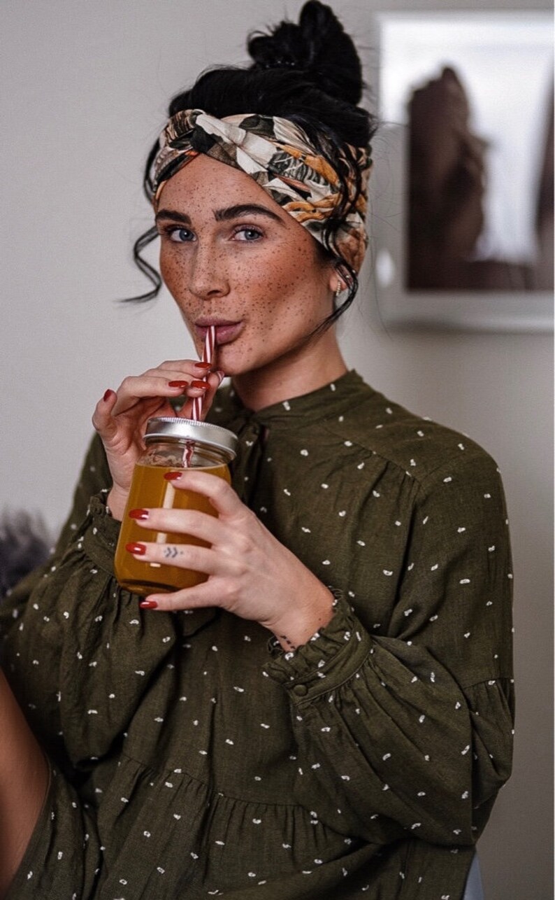 Op de afbeelding: Een vrouw met een patroon hoofdband en een groene blouse met witte stippen drinkt uit een glazen pot met een rietje. De pot bevat een oranje vloeistof. De vrouw heeft rode nagellak.