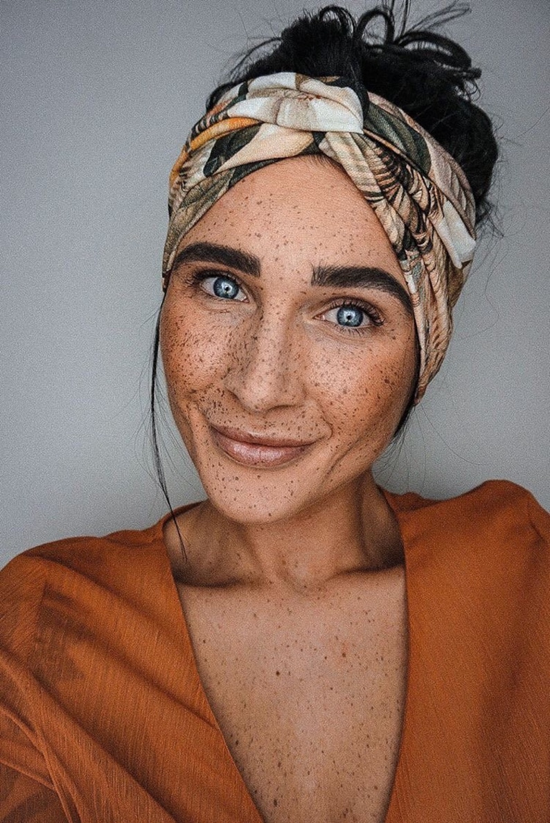 Op de afbeelding: Een vrouw met blauwe ogen en sproeten draagt een hoofdband met bloemenpatroon en een oranje top. Haar donkere haar is opgestoken. De afbeelding is een close-up op een neutrale achtergrond.