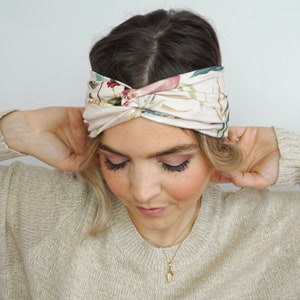 K&ouml;nnte beinhalten: Ein Haarband mit floralem Muster und Knoten-Design. Das Haarband hat einen cremefarbenen Hintergrund mit rosa, gr&uuml;nen und goldenen botanischen Mustern. Das Accessoire wird auf dem Kopf getragen.